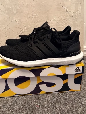 Adidas Ultra Boost svarta sneakers - Svarta Adidas Ultra Boost sneakers med stickad ovandel och vita Boost-sulor. Skorna har klassiska tre ränder på sidan, svart hälkappa med 'ultra boost'-text och elastiska svarta snören. Perfekta för dig som gillar sportig och clean stil.