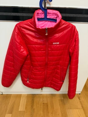 Röd pufferjacka  - Snygg röd pufferjacka med quiltad design och hög krage. Jackan har dragkedja framtill, två sidofickor med dragkedja och en kontrasterande ljusrosa insida. Perfekt för kalla vinterdagar och riktigt skön att bära.