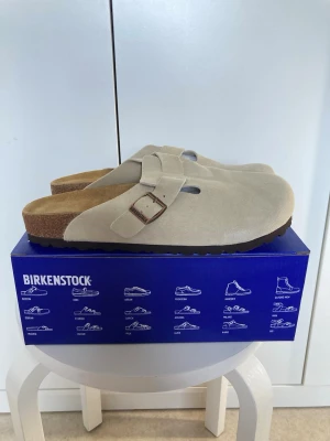 Birkenstock Boston beige tofflor - Säljer ett par Birkenstock Boston tofflor i beige mocka med justerbart spänne i metall. Klassisk slip-in modell med mjuk fotbädd och brun sula. Perfekta för chill dagar hemma eller när du vill ha det bekvämt och stilrent.