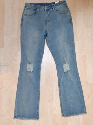 Ljusblå bootcut jeans med slitningar Y2K Skate Japan stil  - Säljer ett par ljusblå bootcut jeans med slitna detaljer på knäna och råa kanter vid bensluten. Jeansen har klassisk femficksmodell och hög midja. Perfekta för en avslappnad och trendig look.