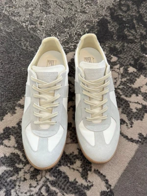 Maison margiela vit  - Snygga sneakers i vitt och ljusgrått med ovandel i läder och mocka. Skorna har klassisk snörning, beige sula och stilren design. Perfekta för dig som gillar minimalistisk och trendig stil.