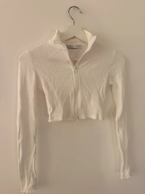 Vit ribbad cropped ziptröja - Trendig vit ribbad ziptröja med lång ärm och hög krage. Croppad modell som ger en snygg siluett. Tillverkad i stretchigt material för en bekväm passform. Perfekt att matcha med höga jeans eller kjol.