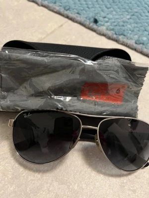Ray-Ban aviator solglasögon  - Ray-Ban solglasögon med silverfärgad metallram och svarta skalmar. Köpta från synoptik för många år sen, men knappt använt. Svart-gråa glas Råkade tappa glasögonen när jag skulle fota så vänstra glaset har fått en  vädligtminimal repa på insidan som knappt går att se Kommer med original fodral och putsduk. Jag är kvinna men tror att glasögonen passar för både könen. 