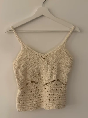 Beige virkad linnetopp - Snygg beige virkad linnetopp med smala axelband och v-ringad hals. Toppen har ett unikt mönster och är perfekt för varma dagar. Passar till både jeans och kjol för en trendig look.