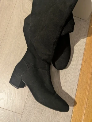 Svarta knähöga stövlar med klack - Snygga svarta knähöga stövlar i mockaimitation med bred klack och rundad tå. Dragkedja på insidan för enkel påtagning. Perfekta till höst- och vinterlooken och passar till både jeans och kjol.