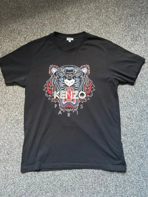 Svart Kenzo t-shirt med tigertryck - Svart t-shirt från Kenzo med ikoniskt tigertryck i blått, rött och vitt framtill samt texten 'KENZO PARIS'. Klassisk passform och rund halsringning. Perfekt för dig som vill sticka ut med en streetstyle-look.