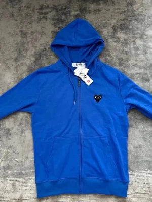 Blå hoodie med dragkedja och hjärtdetalj - Snygg blå hoodie med dragkedja framtill och huva. På bröstet finns en svart hjärtdetalj med ögon, vilket ger en unik och trendig look. Hoodien har fickor på sidorna och ribbade muddar. Perfekt för dig som vill ha en stilren men ändå lekfull hoodie.
