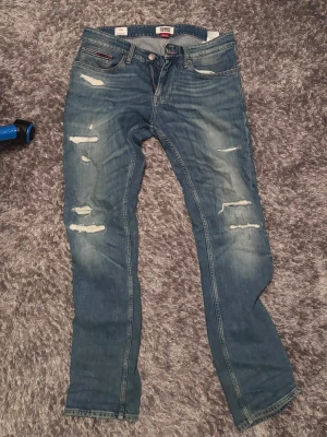 Tommy Hilfiger Jeans  - Säljer ett par blå jeans från Tommy Hilfiger med slitningar och distressed detaljer på benen. Klassisk femficksmodell med smal passform och snygga sömmar. Perfekta för en avslappnad och trendig look.