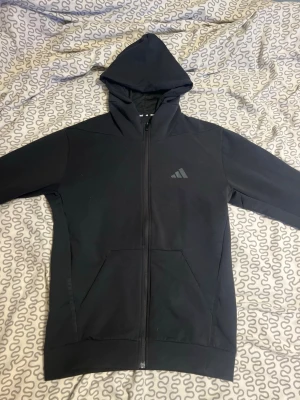 Svart hoodie med dragkedja från Adidas - Säljer en svart hoodie från Adidas med hel dragkedja framtill och två stora fickor. Klassisk Adidas-logga på bröstet och huva med snörning. Perfekt för chill dagar eller när du vill ha en enkel och stilren look.