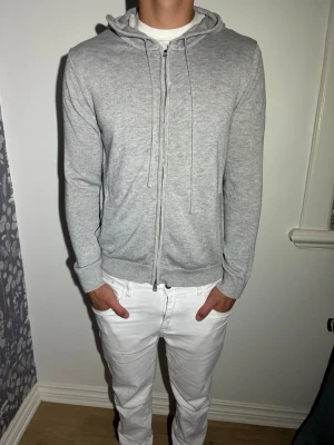 Ljusgrå Kashmirzip hoodie med huva - Snygg ljusgrå hoodie med dragkedja och huva. Tillverkad i mjukt material med ribbade muddar vid ärmar och nederkant. Klassisk passform och dragsko i huvan för justerbar stil. Perfekt till jeans eller joggers för en chill look. Han på bilden är 180 och väger 79 kg