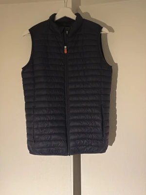 Mörkblå quiltad dunväst av save the duck - Snygg mörkblå dunväst från save the duck med quiltat mönster och hög krage. Västen har dragkedja framtill med en liten orange detalj på dragkedjan. Perfekt att slänga över en hoodie eller tröja för extra värme. Klassisk och stilren design utan ärmar. Finns inga tecken på defekter.
