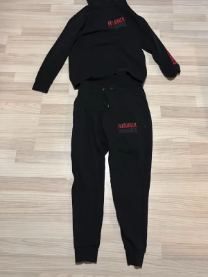 Svart Hoodrich loungewear set - Säljer ett svart loungewear set från Hoodrich med matchande hoodie och byxor. Båda plaggen har Hoodrich-logga i rött och vitt tryck på bröstet och benet. Hoodien har huva och byxorna har dragsko i midjan samt muddar vid benslut. Perfekt för chill dagar.