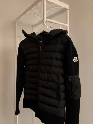 Moncler Cardigan Svart - Snygg svart cardigan från Moncler med quiltad front och rygg i glansigt material samt stickade ärmar och sidor. Jackan har huva, dragkedja framtill, fickor med dragkedja och Moncler-logga på ärmen. Perfekt nu till hösten🍂 Skriv till mig vid minsta frågor eller funderingar😊