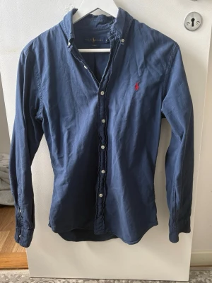 Skjorta Ralph lauren  - Klassisk marinblå skjorta från Ralph Lauren i slim fit-modell. Skjortan har långa ärmar, knappar framtill och den ikoniska röda logotypen broderad på bröstet. Tillverkad i bomull för en skön känsla och stilren look.