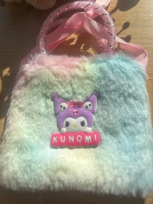 Pastellfärgad fluffig small handväska - Supersöt handväska i pastellfärgad fluff med motiv av Kunomi och texten 'KUNOMI' på framsidan. Väskan har rosa handtag med glittriga detaljer och en stor rosa rosett. Perfekt för dig som gillar kawaii-stil och vill sticka ut. 