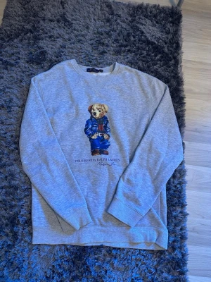 Grå sweatshirt Polo Bear Ralph Lauren - Säljer en grå sweatshirt från Ralph Lauren med den ikoniska Polo Bear-trycket framtill. Tröjan har rund halsringning, långa ärmar och ribbade muddar. Materialet är mjukt och bekvämt, perfekt för en avslappnad stil. Klassisk och tidlös design med snygg detalj framtill.