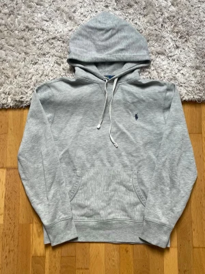 Ljusgrå hoodie med broderad logga - Snygg ljusgrå hoodie med huva och dragsnören. Klassisk känguruficka framtill och en liten broderad logga i mörkblått på bröstet. Tillverkad i mjukt sweatshirtmaterial som är perfekt för chill dagar. Passar dig som gillar enkel och stilren streetwear. Har en pytte liten röd fläck som inte riktigt syns men vill ändå påpeka. Se i bild 