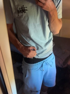 Ljusblått Stüssy set med tryck - Säljer ett ljusblått set från Stüssy med t-shirt och shorts. T-shirten har Stüssy-logga framtill och ett stort tärningsmotiv med logga på ryggen. Shortsen har resår i midjan, dragsko och logga på ena benet. Perfekt för en avslappnad streetwear-look.