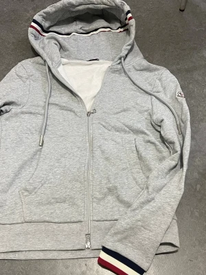 Grå zip-hoodie från Moncler - Snygg grå hoodie med dragkedja från Moncler. Huva med dragsnören och coola röd-blå-vita detaljer på insidan av huvan och ärmslut. Mjukt material och två fickor framtill. Perfekt för en avslappnad och trendig look.