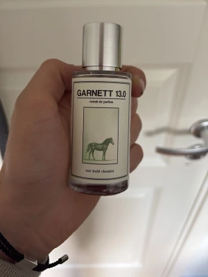 GARNETT 13.0 extrait de parfum - Herr parfymflaska med stilren design och silverfärgat lock. Etiketten har en grön häst och texten 'one bold chemist'. Flaskan är genomskinlig och cylindrisk, vilket ger en modern och minimalistisk känsla. Perfekt för dig som gillar unika dofter.