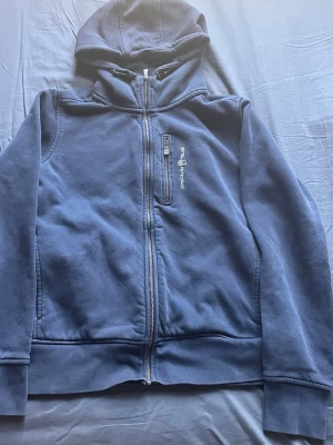 Marinblå hoodie från Sail Racing - Snygg marinblå hoodie från Sail Racing med dragkedja framtill och en extra bröstficka med dragkedja. Broderad logga på bröstet och huva med dragsko. Tillverkad i mjukt bomullsmaterial, perfekt för chill dagar eller när du vill ha en clean look.