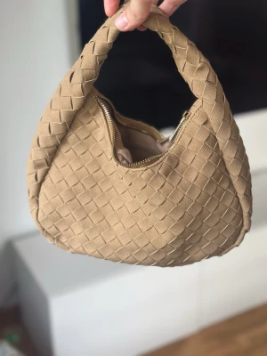 Beige flätad handväska - Trendig beige handväska med flätad design och rundad form. Väskan har ett kort handtag och stängs med dragkedja. Perfekt accessoar för att lyfta din outfit med en stilren och modern touch