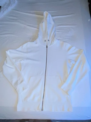 Vit zip hoodie med huva - Säljer en stilren vit hoodie med dragkedja framtill och stor huva. Hoodien har två fickor på magen och ribbade muddar vid ärmar och nederkant. Perfekt för dig som gillar en clean och enkel look. Materialet känns mjukt och bekvämt.
