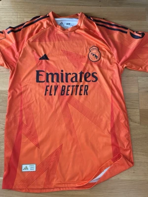 Orange Real Madrid målvaktströja - Säljer en orange Real Madrid målvaktströja från Adidas med svarta detaljer och tryck. Tröjan har klubbmärke och sponsorlogga på bröstet samt Mbappé och nummer 6 på ryggen. Tillverkad i lätt och ventilerande funktionsmaterial, perfekt för fotboll.