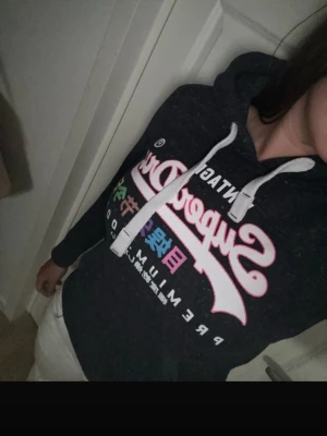 Svart/mörkgrå Superdry hoodie med tryck - Säljer en mörkgrå hoodie från Superdry med färgglatt tryck och text på bröstet. Hoodien har vit dragsko i huvan och ribbade muddar vid ärmslut och nederkant. Perfekt för en avslappnad och cool stil.