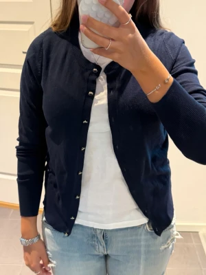 Mörkblå kofta från Holly & White - Klassisk mörkblå kofta från Holly & White med rund halsringning och pärlemorskimrande knappar framtill. Långärmad modell i mjukt material som passar perfekt till lager på lager. Enkel och stilren design med liten logodetalj vid nederkanten.
