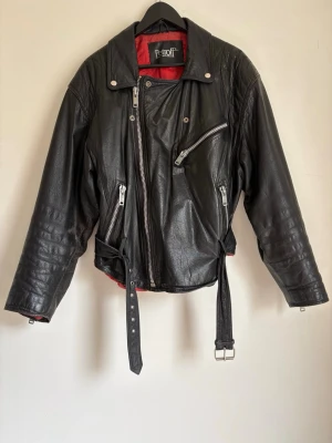 Svart skinnjacka med röd insida - Cool svart skinnjacka från Petroff med röd quiltad insida, sneddragna silverfärgade dragkedjor, bälte i midjan och klassisk bikerstil. Jackan har flera fickor och tryckknappar vid kragen för extra edge. Välanvänd men vintage😎