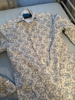Vit blommig skjorta med blått mönster - Snygg vit skjorta med blått blommigt mönster. Skjortan har långa ärmar, klassisk krage och knäppning framtill. Perfekt för dig som gillar stilrena men ändå unika plagg. Tillverkad i mjukt bomullstyg för skön känsla.