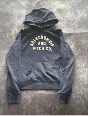 Mörkblå hoodie Abercrombie & Fitch - Mörkblå hoodie från Abercrombie & Fitch med stor vit logga på bröstet. Klassisk känguruficka framtill och ribbade muddar vid ärmslut och nederkant. Mjuk bomullskänsla och avslappnad passform. Perfekt för en chill och casual look. Storlek L men passar bättre på M.