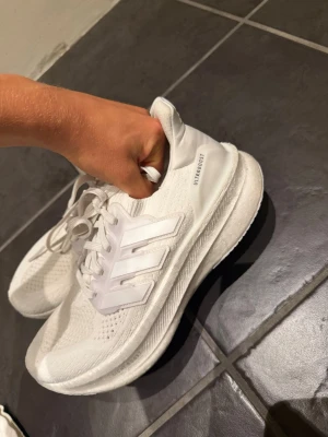 Vita Adidas Ultraboost sneakers - Säljer ett par helvita Adidas Ultraboost sneakers med klassiska tre ränder på sidan. Skorna har en stickad ovandel i mesh för bra andningsförmåga och en tjock, bekväm sula. Perfekta för dig som gillar stilrena och sportiga sneakers.