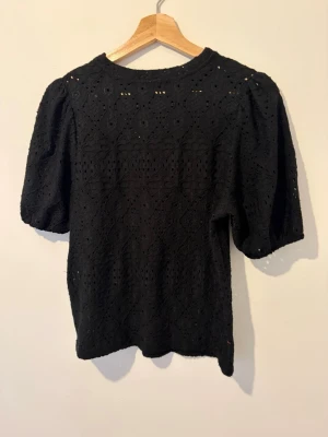 Svart blus med puffärm och spetsdetaljer - Snygg svart blus med puffärmar och vackra spetsdetaljer i ett genombrutet mönster. Blusen har rund halsringning och korta ärmar, perfekt för dig som vill ha en trendig och feminin topp till din outfit.