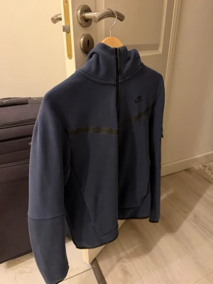 Blå ziptröja från Nike - Mörkblå ziptröja från Nike med hög krage och hel dragkedja framtill. Tröjan har en diskret svart logga på bröstet och en svart rand över bröstet. Tillverkad i mjukt material och har en ficka på ärmen. Inga defekter eller skador! 