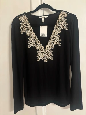 Svart långärmad topp med spetsdetalj - Svart långärmad topp med djup v-ringning dekorerad med beige spets i blommigt mönster. Tunn och mjuk kvalitet som sitter snyggt på kroppen. Perfekt för dig som vill ha något extra till din outfit.