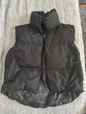 Svart dunväst med hög krage - Snygg svart dunväst med hög krage och dragkedja framtill. Västen har två sidofickor och justerbar dragsko nedtill för extra passform. Perfekt att slänga över en hoodie eller tröja när det är kyligt ute. Klassisk och enkel design som funkar till det mesta.