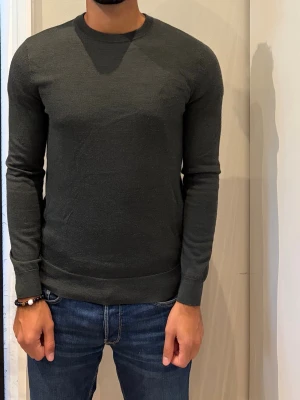 Mörkgrå stickad tröja herr - En stilren mörkgrå stickad tröja med rund hals och långa ärmar. Tröjan har en smal passform och ribbade muddar vid ärmslut och nederkant. Perfekt att matcha med jeans för en clean look.
