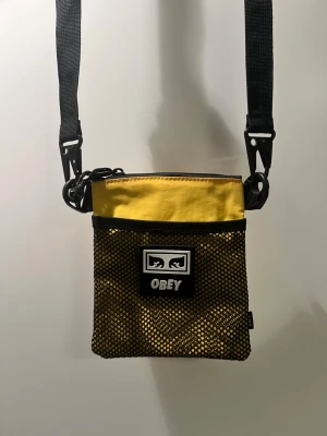 Gul och svart axelväska från OBEY - Snygg gul och svart axelväska från OBEY med justerbar rem och meshficka framtill. Väskan har dragkedja upptill och en tydlig OBEY-logga på framsidan. Perfekt för att bära det viktigaste och ger en streetwear-vibe till din outfit. Det finns en liten fläck på baksidan (bild 3).