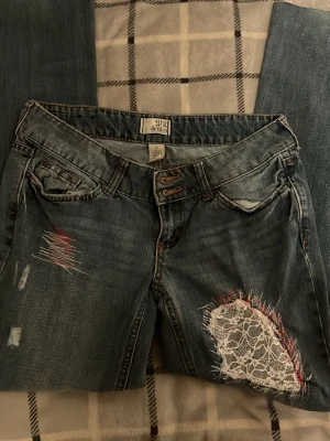 Blå bootcut jeans med spetsdetaljer - Snygga blå bootcut jeans med slitningar och coola detaljer som vit spets och rosa broderier på låret och benet. Jeansen har dubbla knappar i midjan och klassisk femficksmodell. Perfekt för dig som gillar en unik och trendig stil.