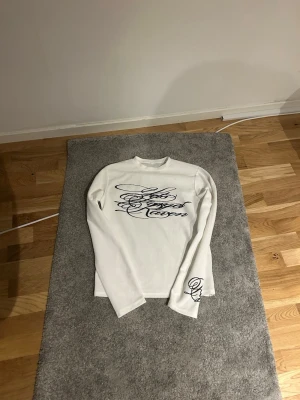 Neverforget långarmad - Säljer en vit långärmad topp med svart tryck i snirklig text över bröstet och på ena ärmen. Toppen är ribbad och har en normal passform med rund hals. Perfekt att styla med jeans.