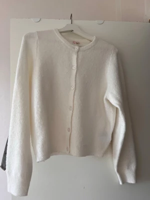 Vit fluffig kofta med rund hals - Mysig vit kofta med rund hals och knappar framtill. Koftan har en fluffig, mjuk struktur och långa ärmar med ribbade muddar. Enkel och stilren design som passar till många olika outfits.