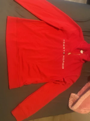 Röd Tommy Hilfiger hoodie med dragkedja - Snygg röd hoodie från Tommy Hilfiger med halv dragkedja och logga tryckt vertikalt på bröstet. Tröjan har långa ärmar, ribbade muddar och är gjord i mjukt sweatshirtmaterial. Perfekt för en avslappnad och trendig look.