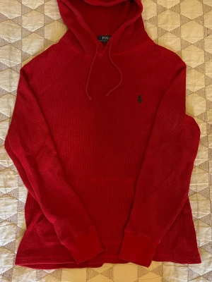 Röd hoodie från Polo Ralph Lauren - Snygg röd hoodie från Polo Ralph Lauren i ett strukturerat, waffle-mönstrat tyg. Klassisk huva med dragsko, magficka och broderad logga på bröstet. Perfekt för dig som vill ha en stilren och bekväm look.