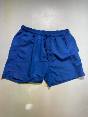 Blå shorts med snörning - Snygga blå shorts med elastisk midja och snörning framtill. Shortsen har en loose passform och är tillverkade i ett lätt syntetmaterial, perfekta för varma dagar. Två sidofickor ger extra funktionalitet.