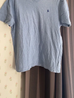 Ljusblå t-shirt med broderad logga - Säljer en ljusblå t-shirt med v-ringning och korta ärmar. T-shirten har en liten broderad logga i mörkblått på bröstet. Tillverkad i mjuk bomull och har en klassisk, normal passform. Perfekt till jeans eller shorts.