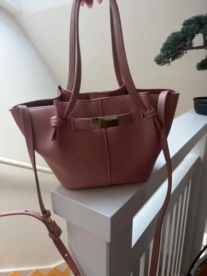 Rosa handväska med gulddetalj - Söt rosa handväska i mjukt skinnimitation med guldfärgad metallspänne framtill. Väskan har både handtag och justerbar axelrem, samt en stilren och modern form. Perfekt för dig som gillar trendiga accessoarer och vill ha plats för det viktigaste.