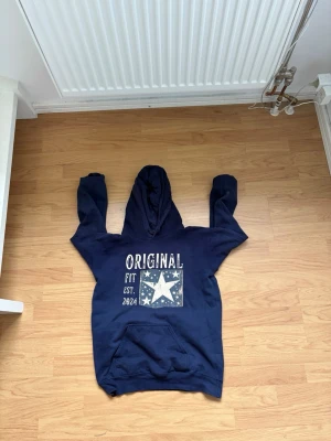 Mörkblå hoodie med stjärntryck - Snygg mörkblå hoodie med stor huva och tryck framtill där det står 'ORIGINAL FIT EST. 2024' tillsammans med en stor vit stjärna och små stjärnor runtom. Hoodien har magficka och ribbade muddar. Perfekt för en chill och avslappnad stil. Limited edition går inte o köpa mer. 