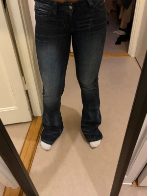 Mörkblå bootcut jeans - Säljer ett par mörkblå bootcut jeans med snygg slitning och klassisk femficksmodell. Jeansen har låg midja och är tillverkade i stretchigt denimtyg som sitter tajt över låren och släpper ut vid bensluten. Perfekta för en avslappnad men trendig look.
